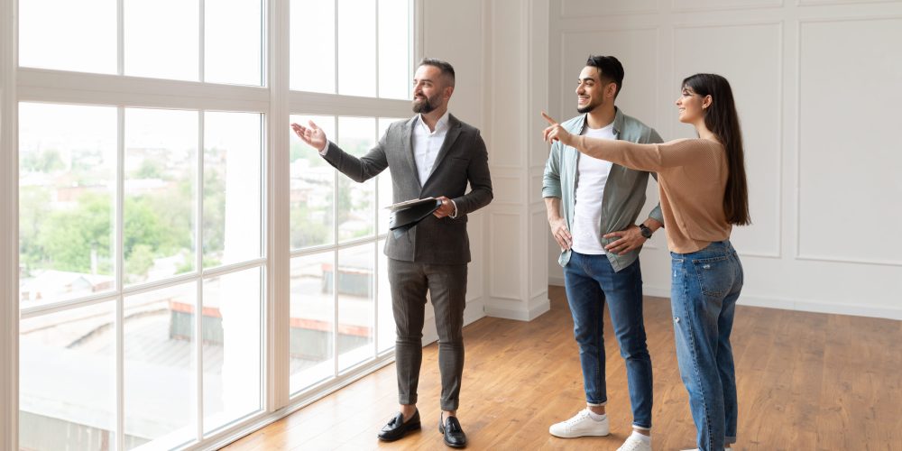 Professional Real Estate Agent Showing Spacious New Apartment To Young Couple, Looking And Pointing At Beautiful View From Window Walls. Happy Middle Eastern Man And Woman Searching For Flat